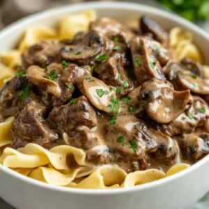 Bowl of Creamy Crockpot Beef Stroganoff over egg noodles, garnished with parsley on a light wooden surface